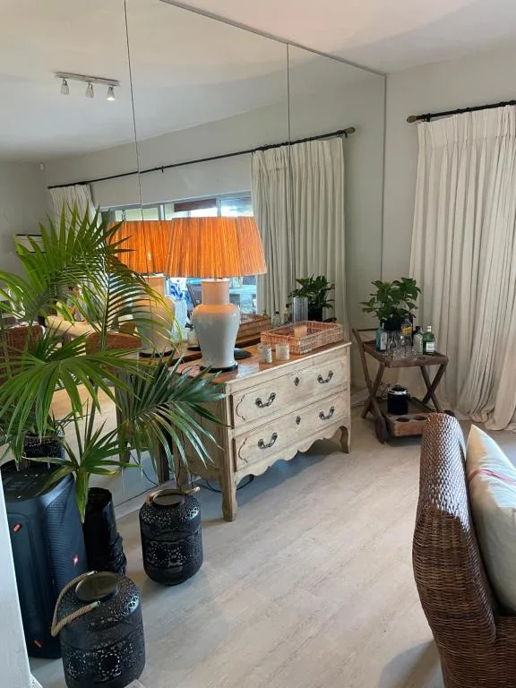 Bright bedroom with large mirror, wooden dresser, potted plants, and cream curtains