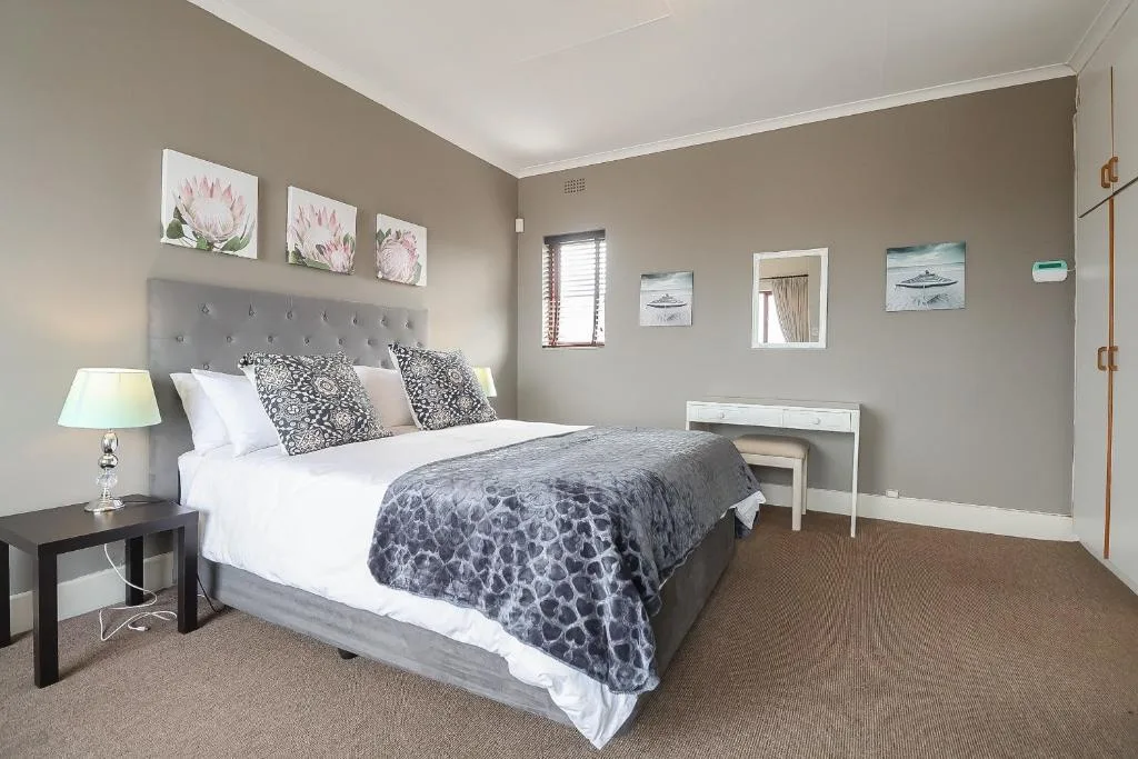 Spacious bedroom with gray upholstered bed, white linens, patterned throw, and coastal artwork