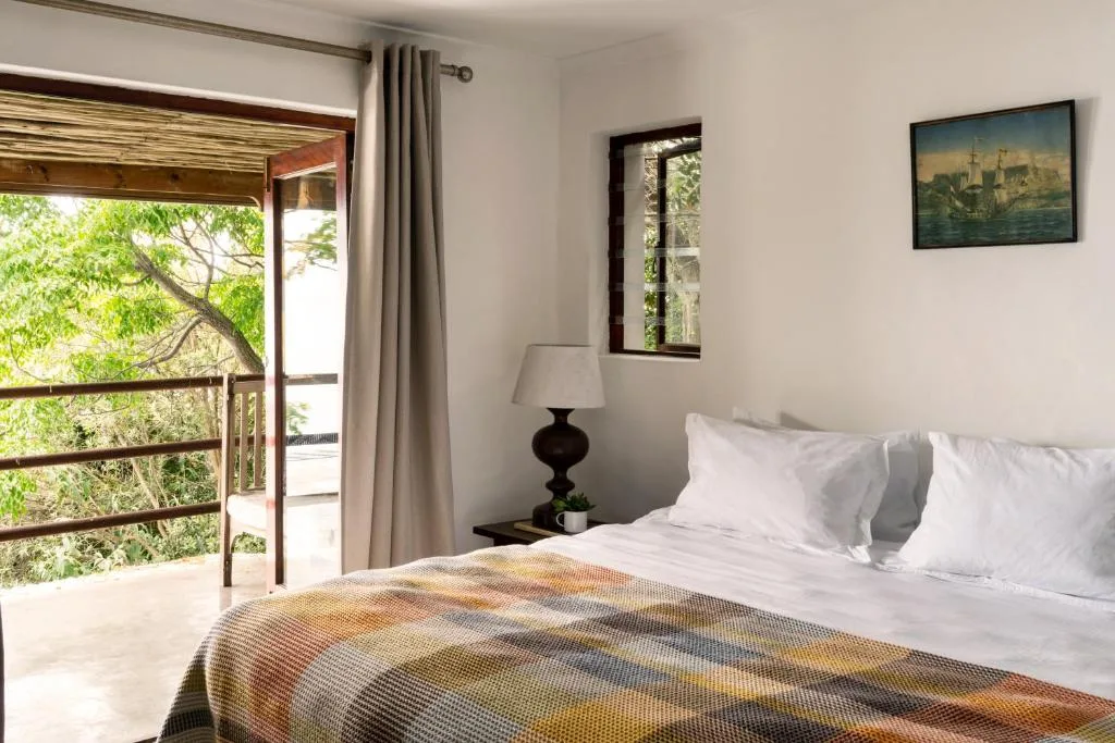Bright bedroom with white pillows, patterned bedspread, and garden view through door