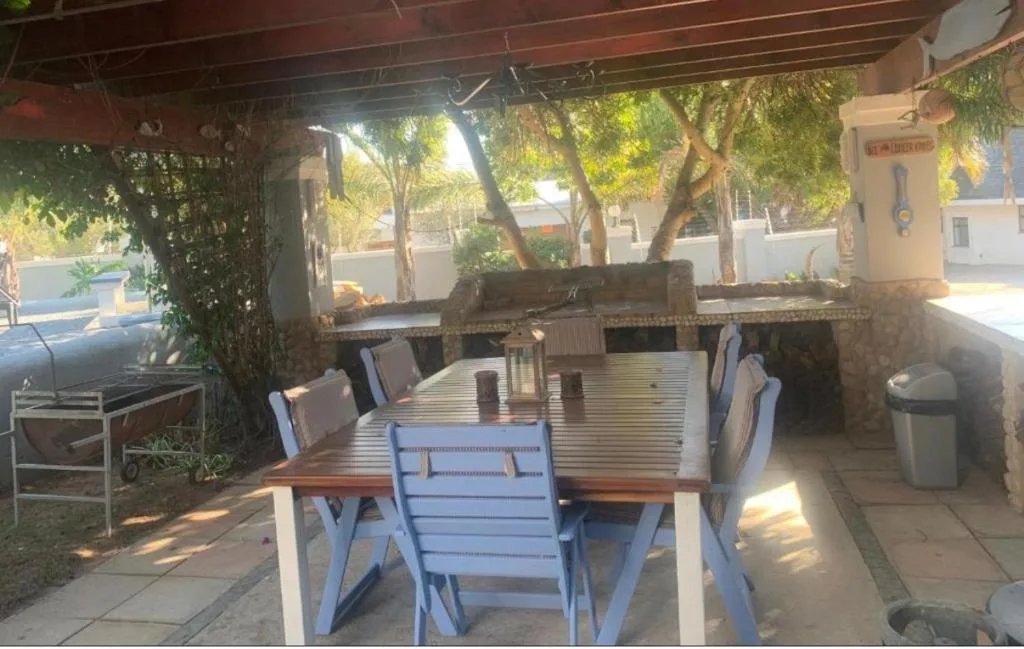 Covered outdoor dining area with wooden table and blue chairs, braai grill, garden setting