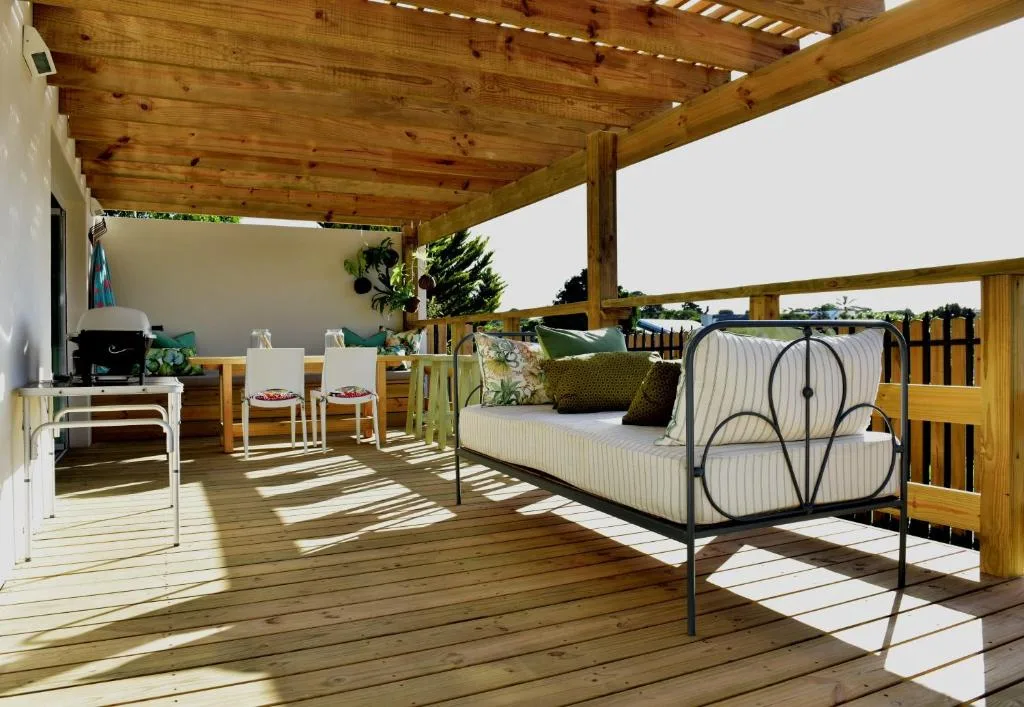 Wooden deck with metal daybed, dining chairs, and braai area under pergola