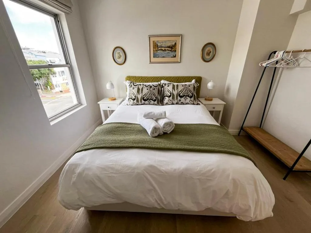 Bright bedroom with olive green headboard and white bedding, townscape visible through window