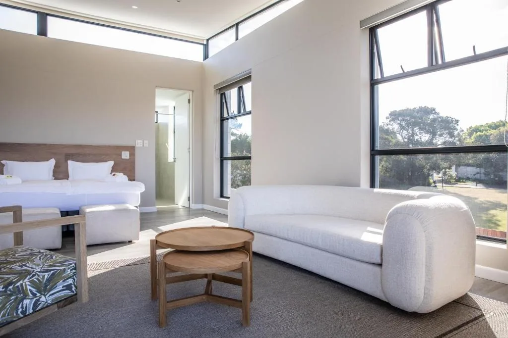 Modern bedroom with bed, white sofa, wooden table, and large windows overlooking trees