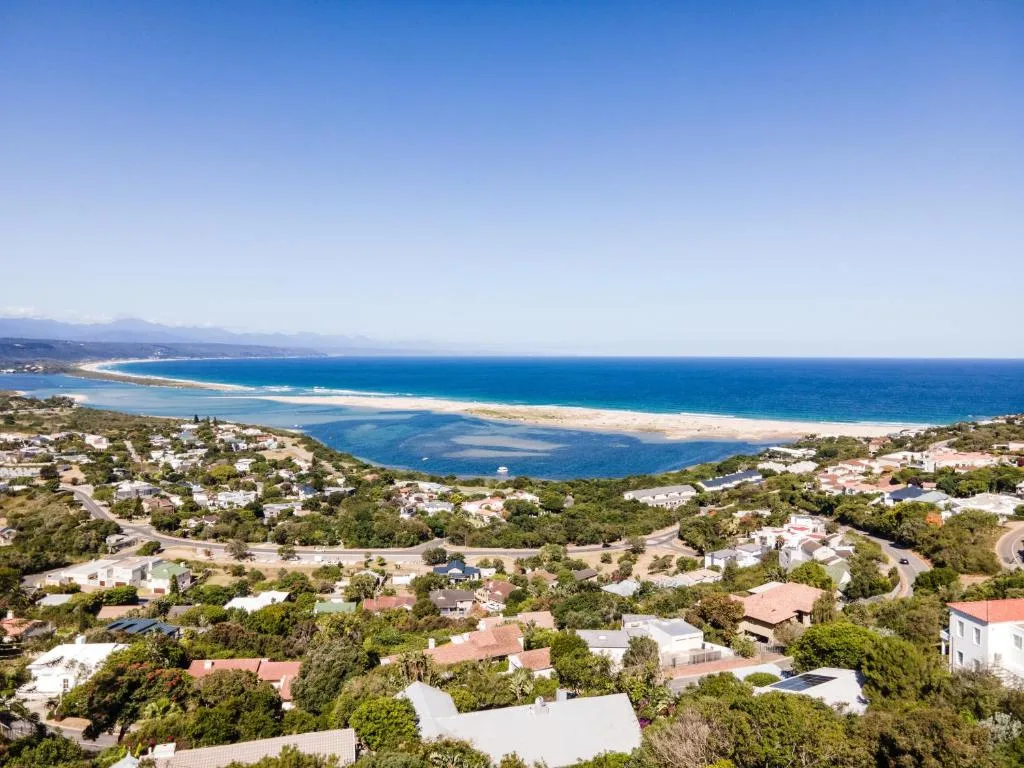 Panoramic coastal view of Plettenberg Bay with golden beach, blue lagoon, and residential properties nestled in lush green hillside landscape