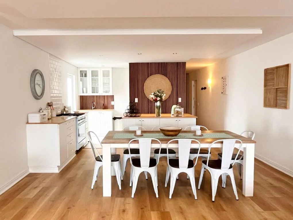 Modern dining area with wooden table and white chairs, open-plan kitchen visible
