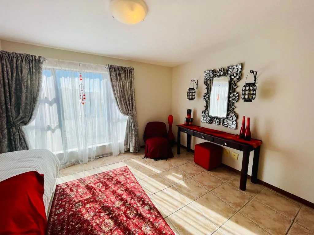 Spacious bedroom with bed, vanity desk, red accents, and large windows overlooking sea