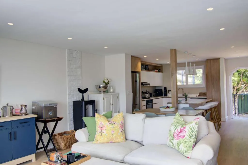 Bright open-plan lounge with cream sofa, colorful cushions, kitchen visible beyond