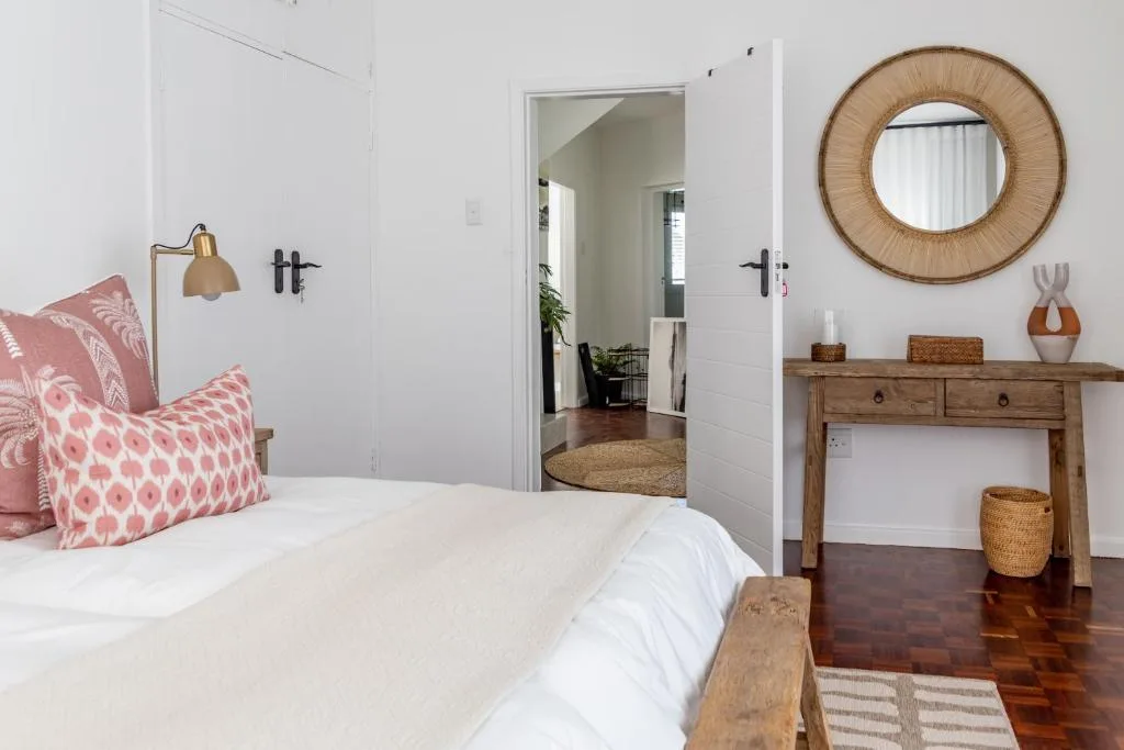 Bright bedroom with white bed, pink patterned pillows, wooden desk, and round mirror