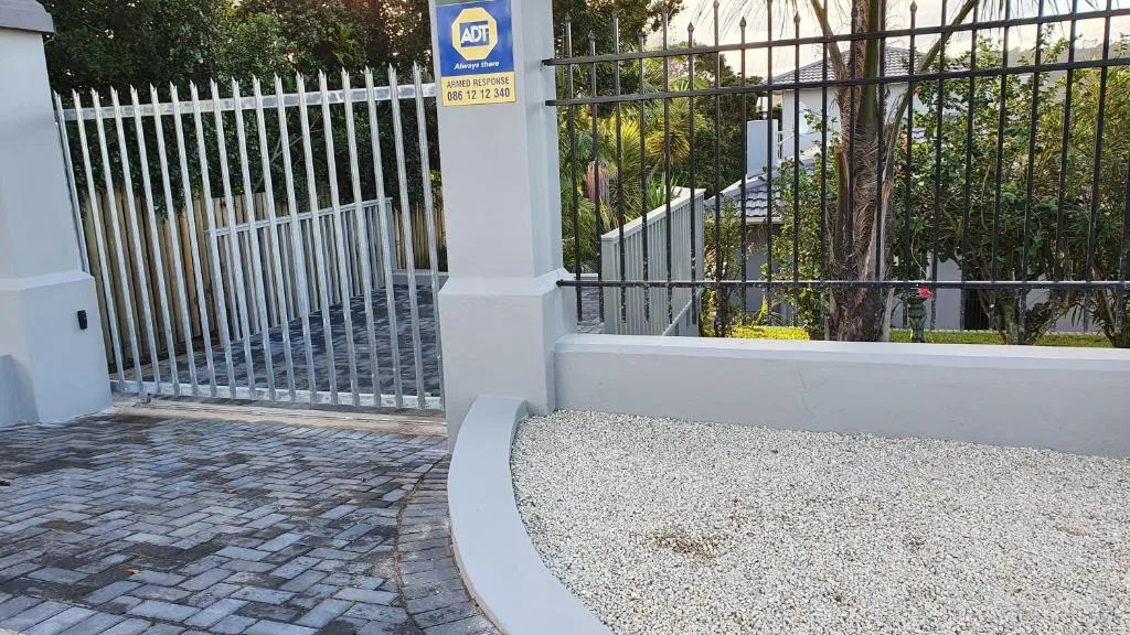 Modern white entrance gate and driveway with security signage and manicured garden
