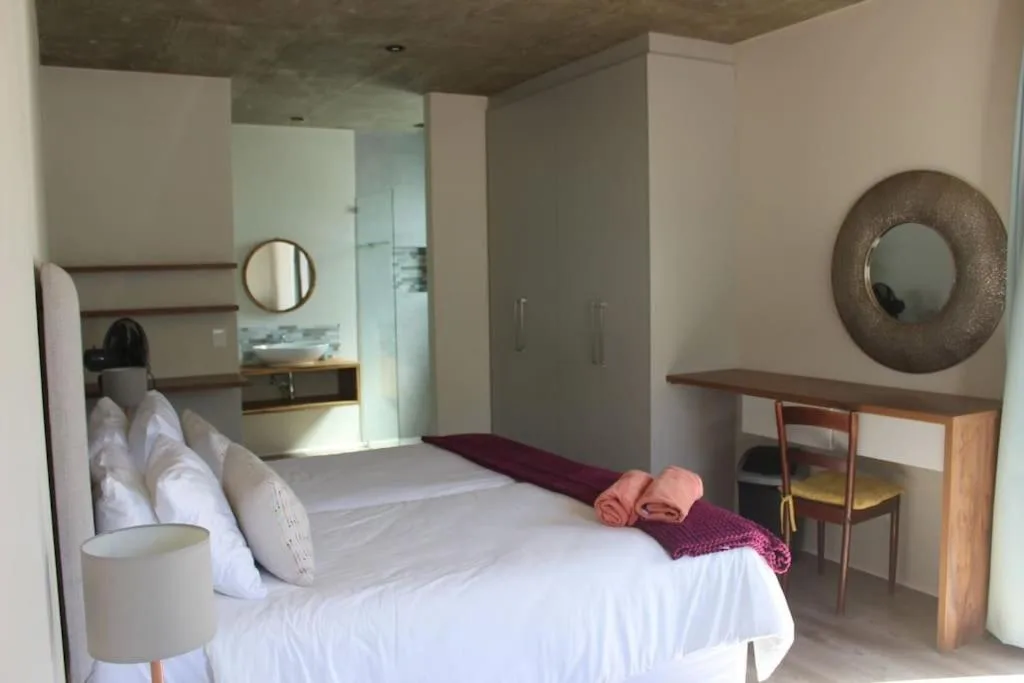 Modern bedroom with white bedding, wooden desk, and en-suite bathroom visible