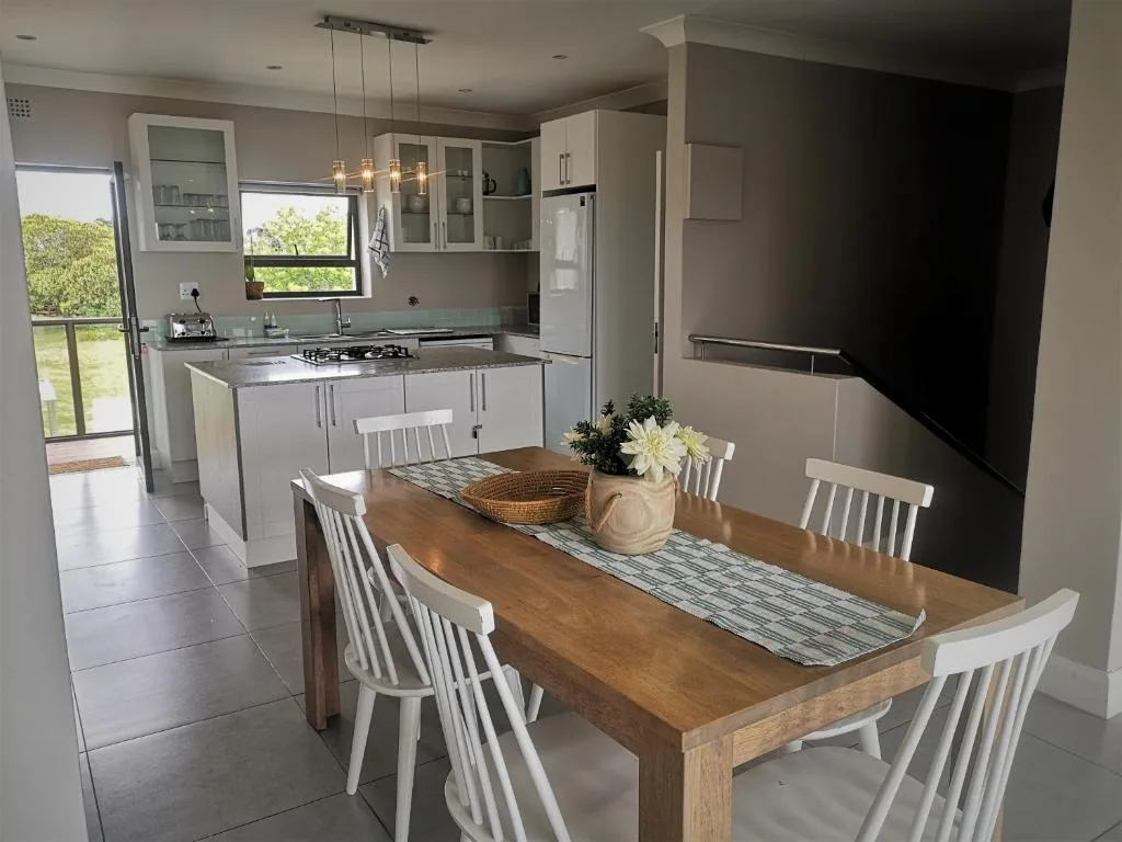 Wooden dining table with white chairs and fresh flowers, kitchen visible beyond