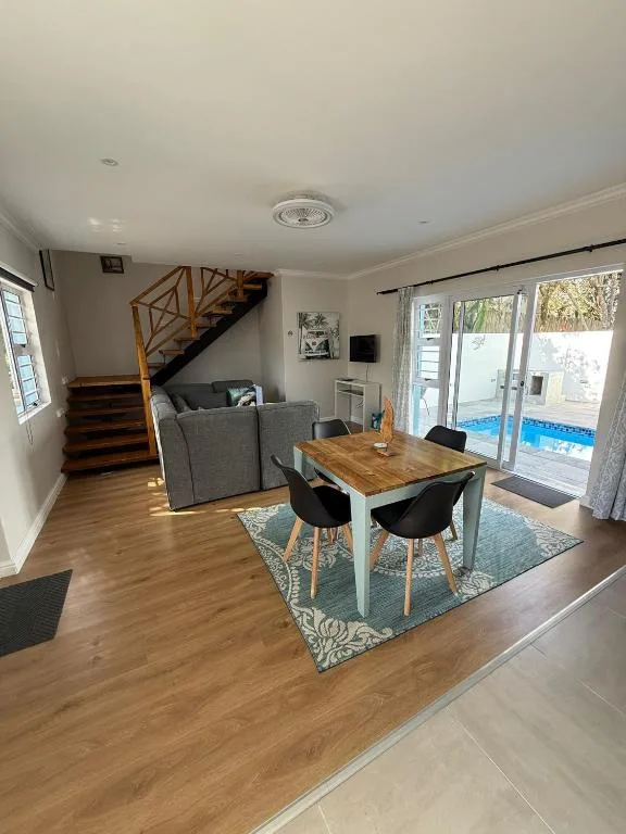 Open-plan lounge and dining area with grey sofa, wooden table, and pool view through glass doors