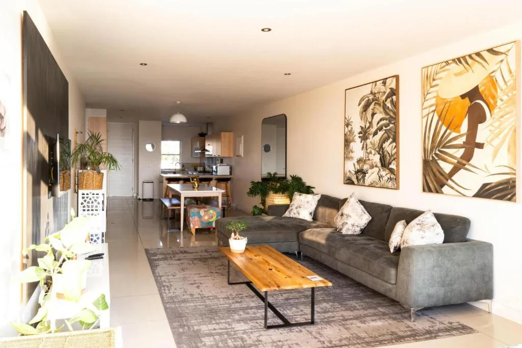 Modern lounge with grey sofa, wooden table, tropical wall art, and open kitchen.