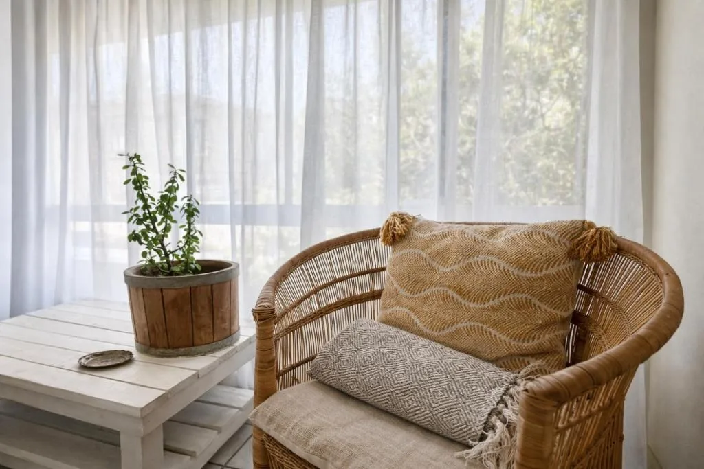Wicker chair with cushions and blanket by sunlit window with sheer curtains