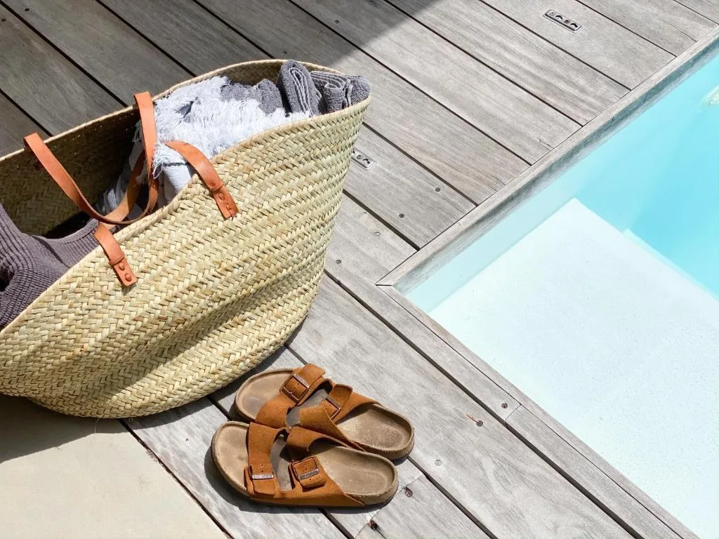 Woven beach basket with towels and sandals on wooden deck