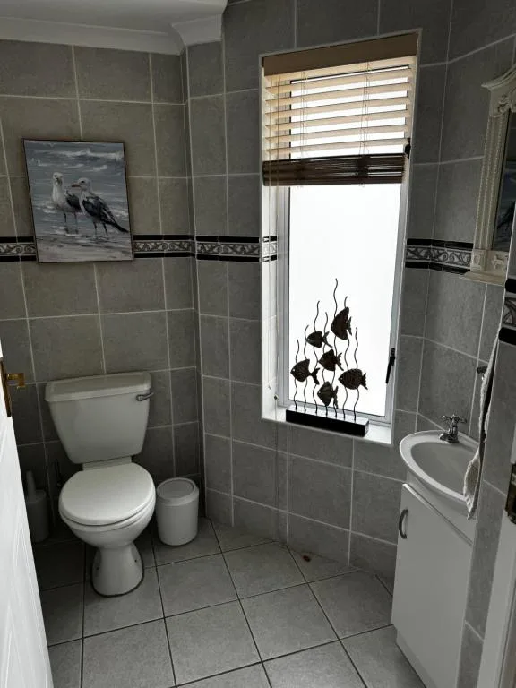 Gray tiled bathroom with toilet, sink, window, and decorative artwork