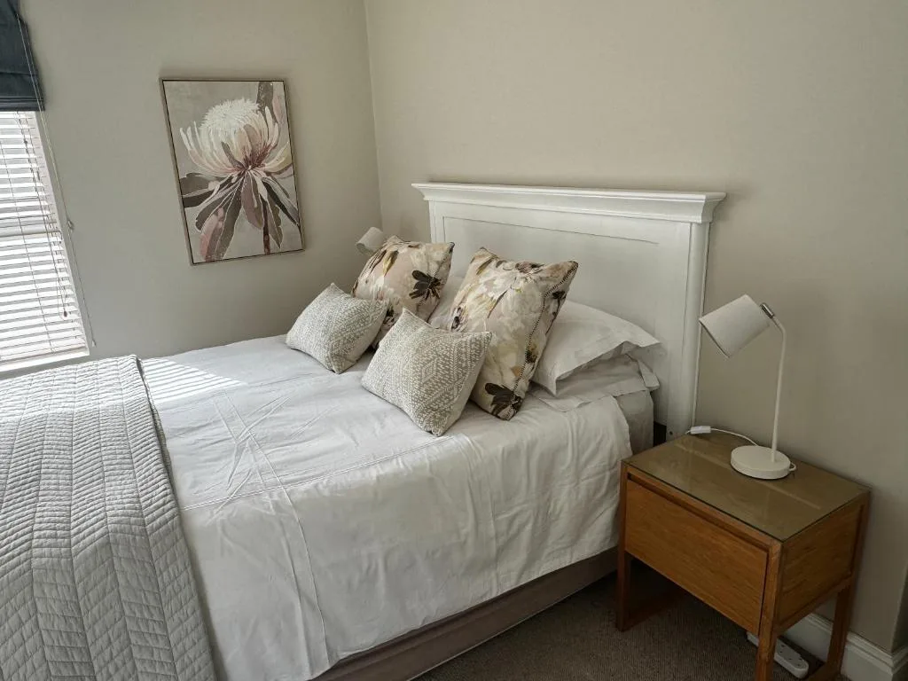 Bright bedroom with white bed, decorative pillows, nightstand, and floral artwork