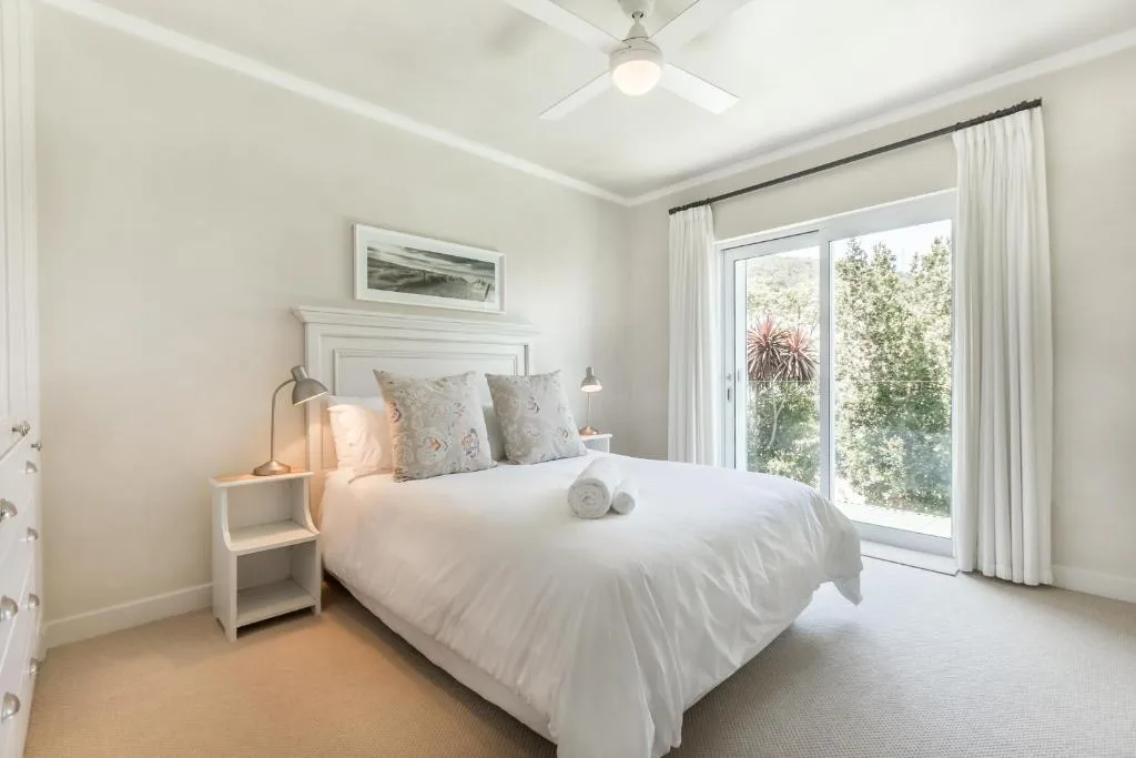 Bright bedroom with white bedding, garden views through glass doors, ceiling fan