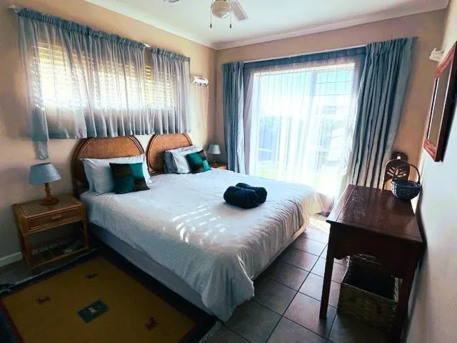 Bright bedroom with queen bed, teal pillows, wooden nightstand, large windows