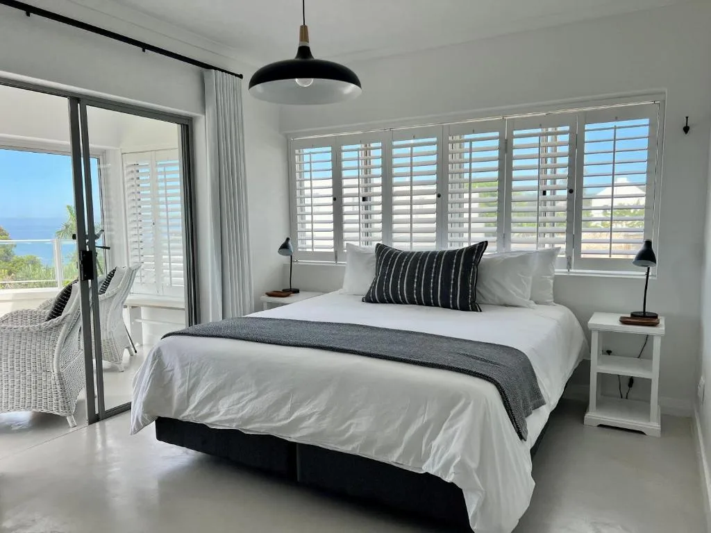Modern bedroom with white bed, ocean view through shuttered windows and glass doors