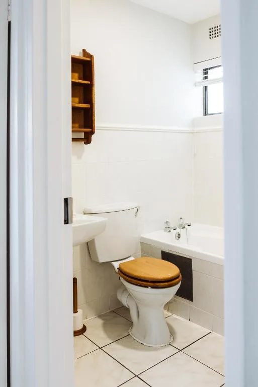 Clean bathroom with white toilet, wooden seat, sink, and wall shelving