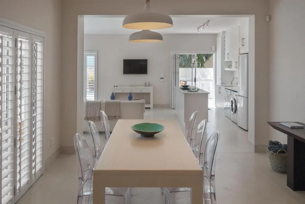 Modern dining area with long table and clear chairs, open-plan kitchen visible