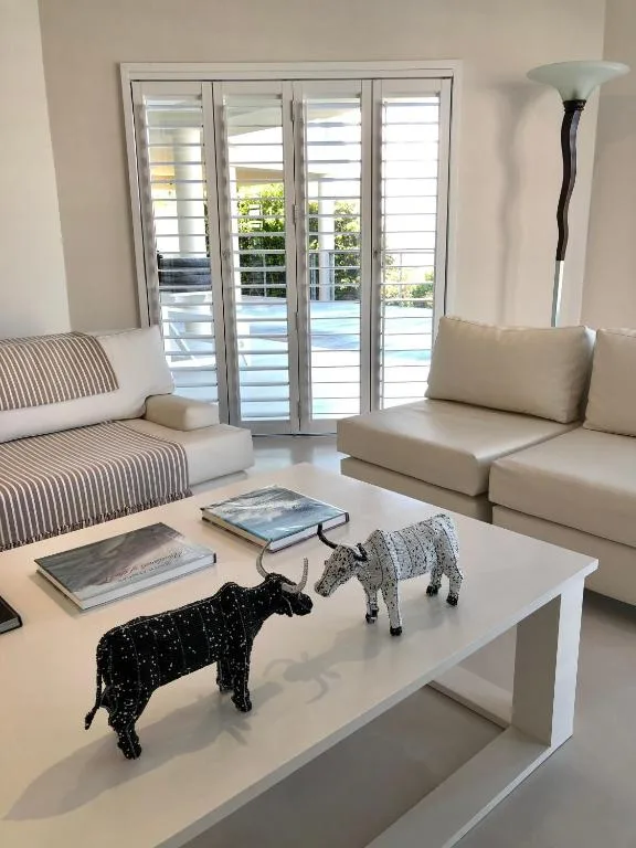 Bright lounge with cream sofas, white coffee table, and shuttered doors opening to pool view
