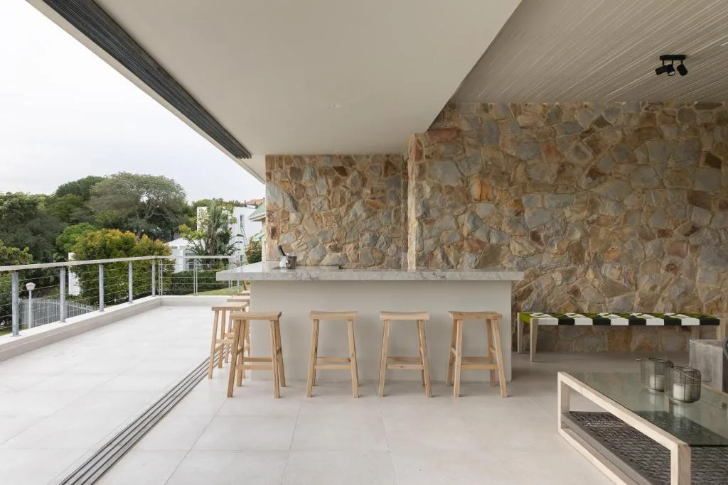 Modern outdoor deck with bar counter, wooden stools, stone wall, and garden views