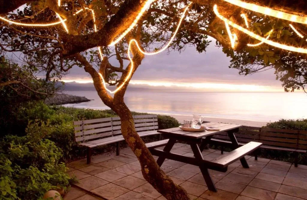 Illuminated tree branches frame outdoor seating area overlooking tranquil lagoon at sunset