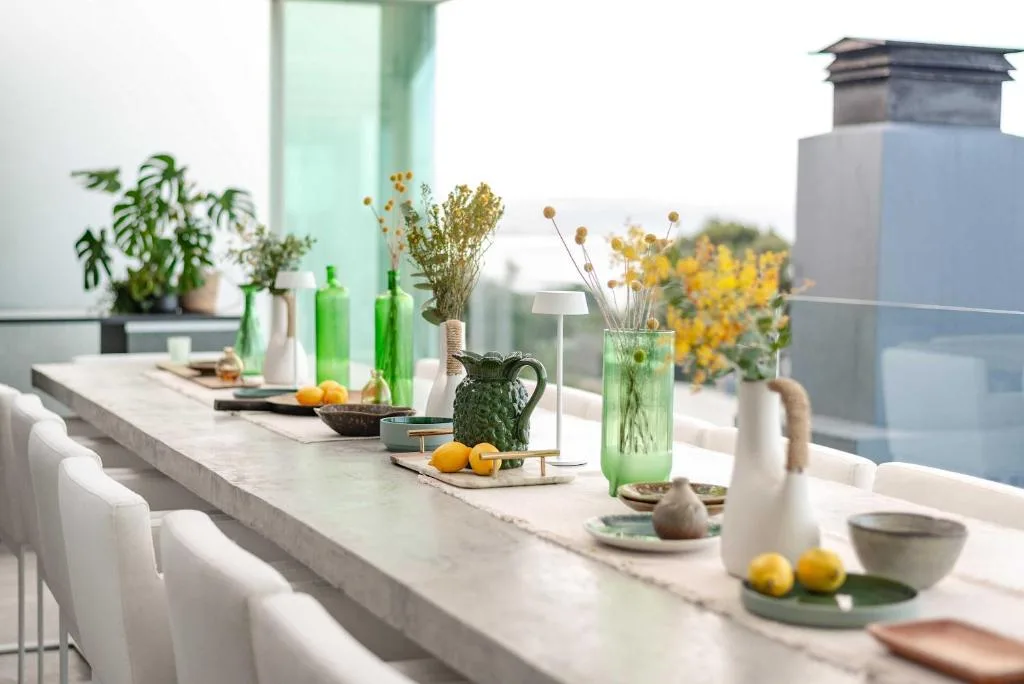 Long marble dining table with white chairs, fresh flowers, and ocean view backdrop