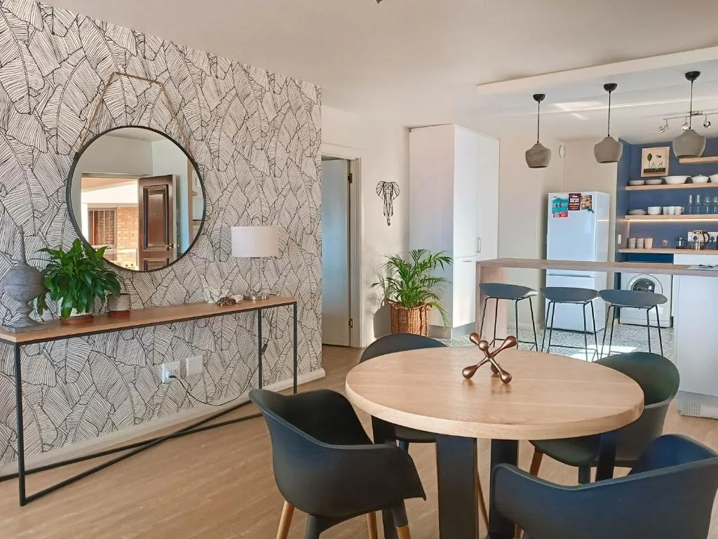 Modern dining area with round wooden table and black chairs, adjacent kitchen visible