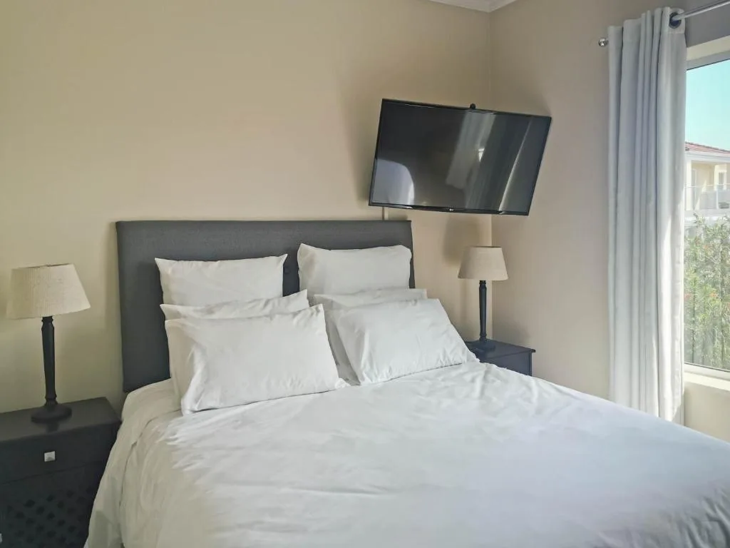 Modern bedroom with white linens, dark headboard, wall-mounted TV, and garden view