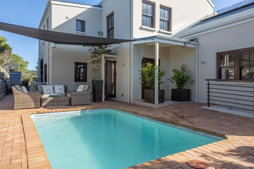 Rectangular swimming pool with turquoise water on brick patio, modern white house with shade sail
