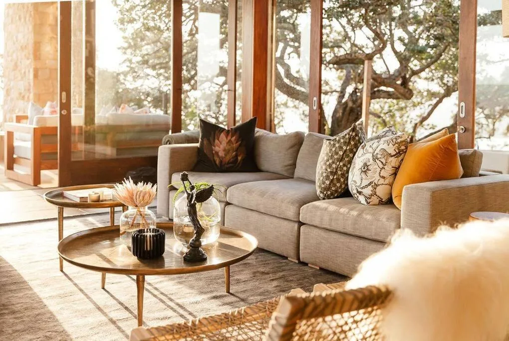 Bright lounge with beige sofa, decorative pillows, and wooden coffee tables overlooking trees
