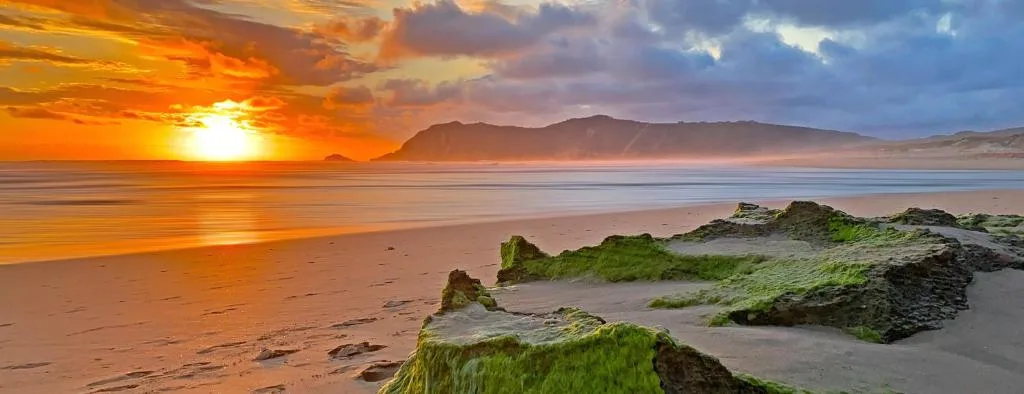 Dramatic sunset over beach with mountains and moss-covered rocks