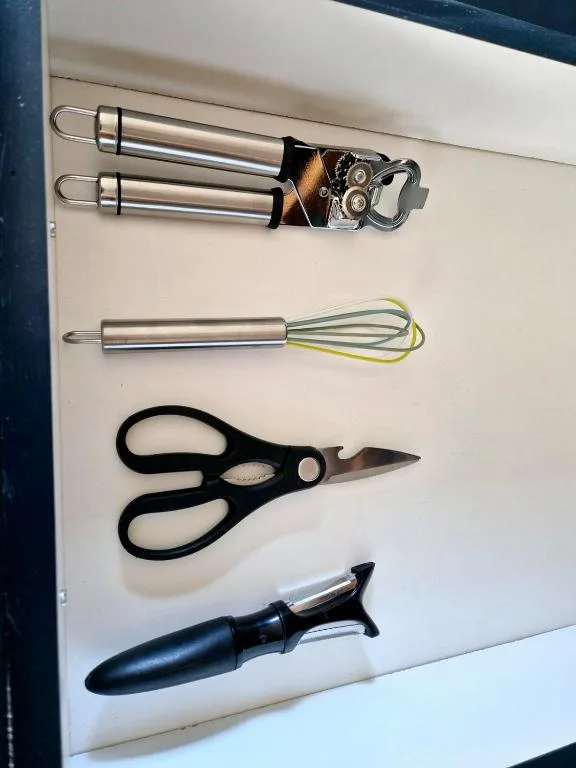 Kitchen utensils including can opener, whisk, scissors, and peeler displayed on white surface