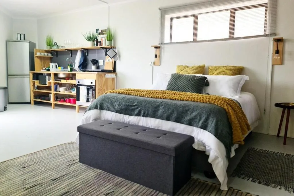 Bright studio bedroom with bed, kitchenette, and yellow accent pillows