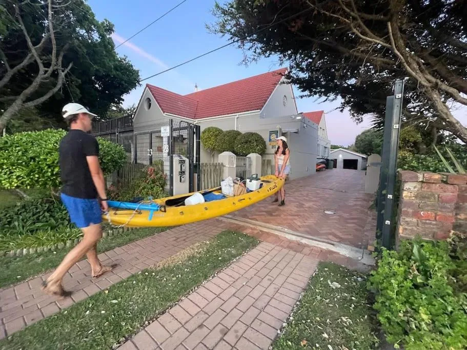 White cottage with red roof, brick driveway, mature trees, kayak storage