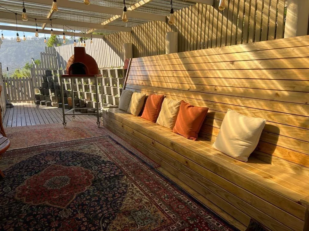 Covered outdoor deck with wooden bench seating, colorful pillows, and braai oven