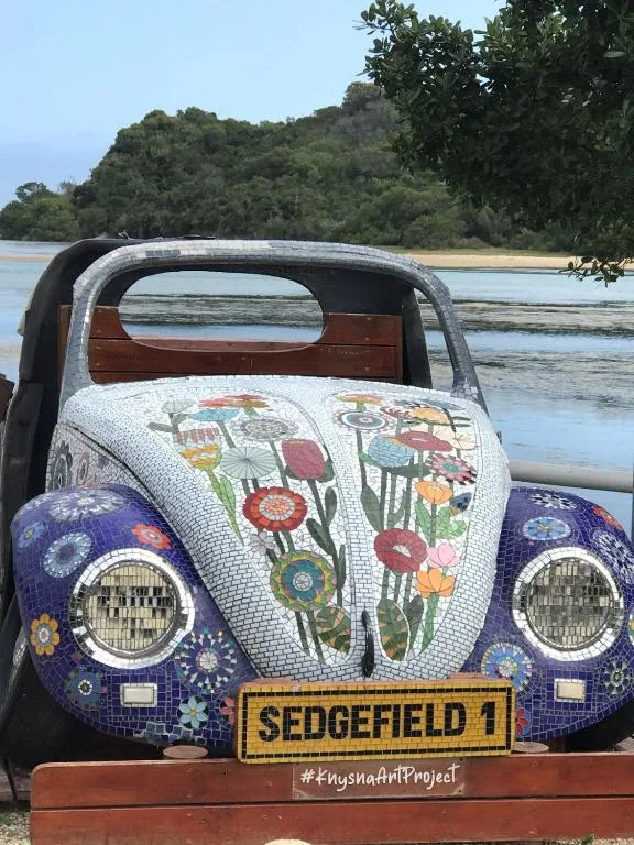 Colorful mosaic Volkswagen Beetle art installation overlooking lagoon and forested hills