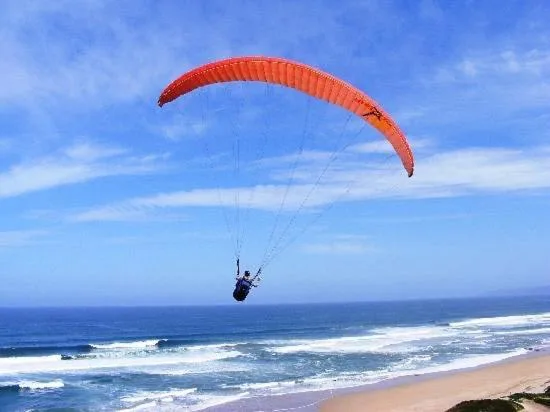 Paraglider with red canopy soaring above pristine beach and ocean waves