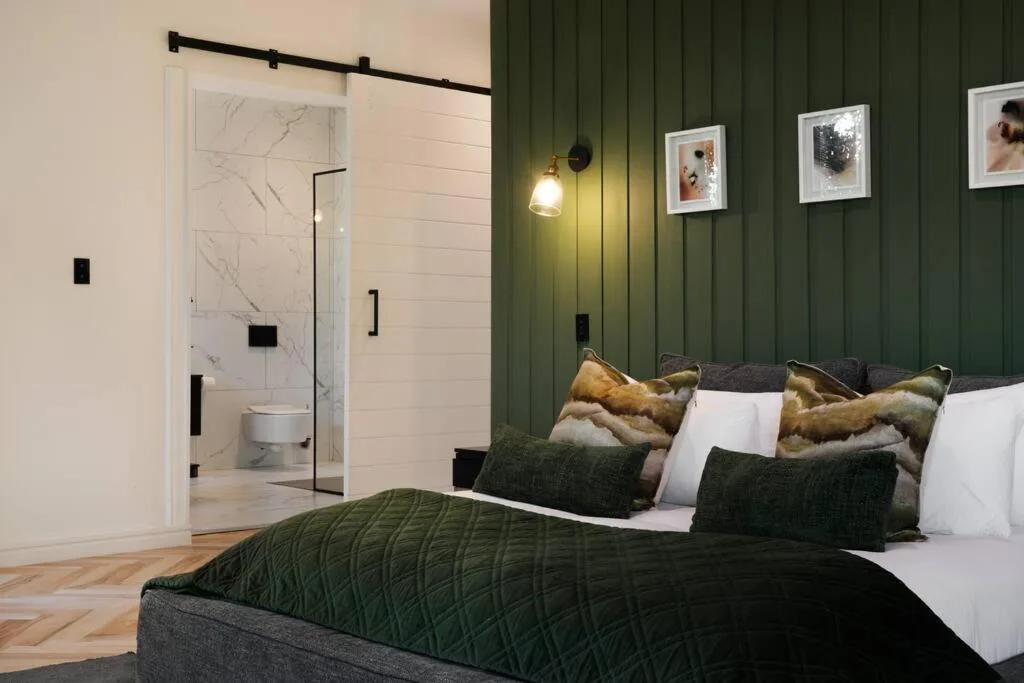 Modern bedroom with green accent wall, bed with gold pillows, and ensuite bathroom