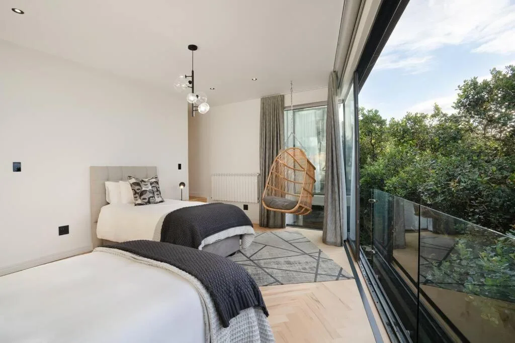 Modern bedroom with twin beds, hanging chair, and garden views through large windows