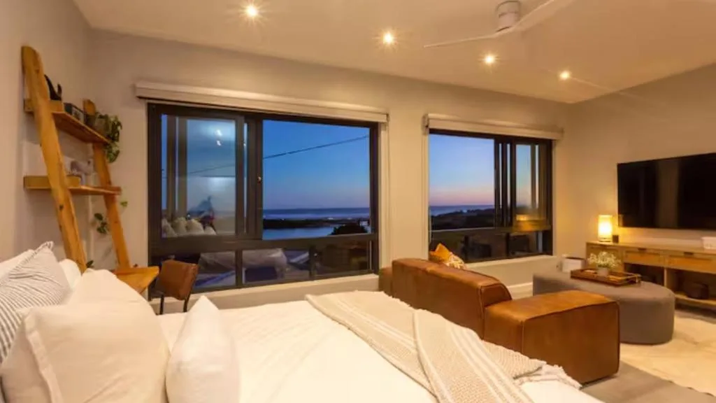Master bedroom with ocean view windows and modern furnishings at dusk