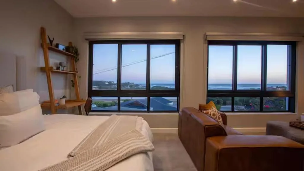 Bedroom with bed, leather seating, and expansive windows overlooking coastal landscape