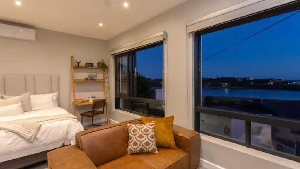Modern bedroom with bed, seating area, and twilight lagoon view through large windows