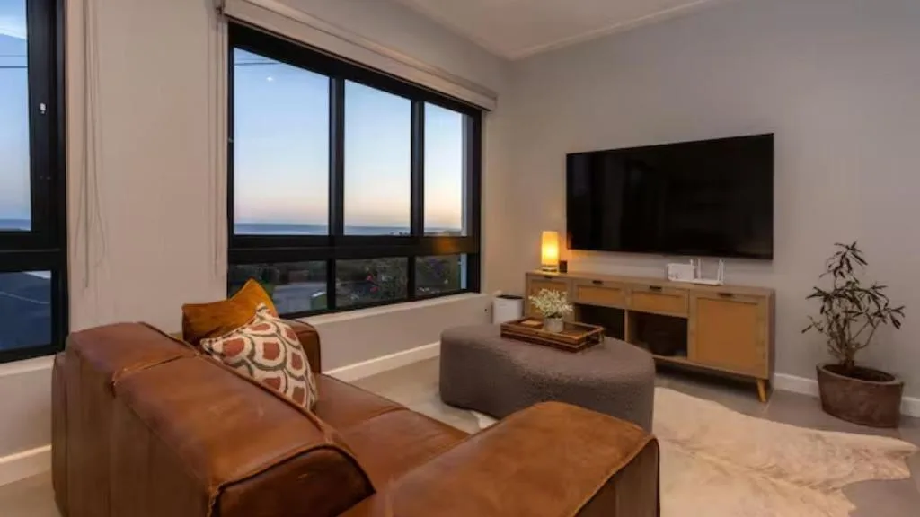 Modern lounge with brown leather sofa, TV, and ocean view windows at dusk