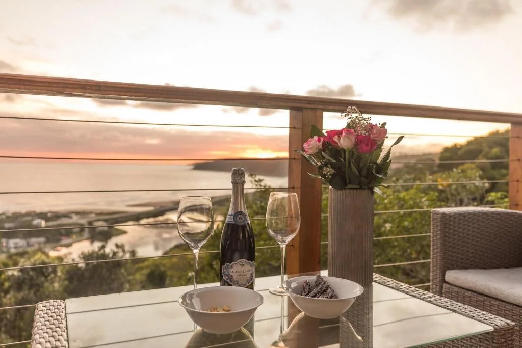 Elevated deck with wine glasses, champagne bottle, and pink flowers overlooking lagoon at sunset