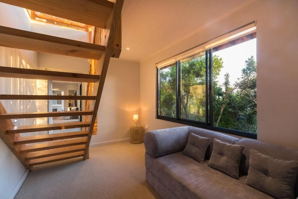 Modern lounge with grey sofa, wooden staircase, and large windows overlooking green landscape