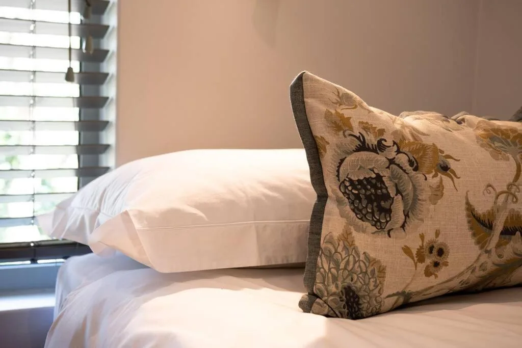 Decorative embroidered pillow on white bedding with window blinds visible